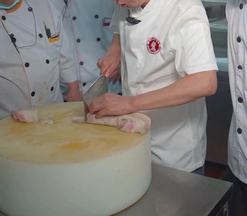 Uno chef usa una mannaia per affettare un blocco di pancetta di maiale bollita su un tagliere grande e spesso.