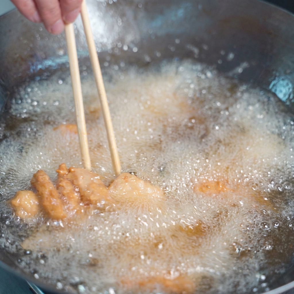 Pezzi di maiale impanati fritti in abbondante olio dorato, mescolati con bacchette di legno.