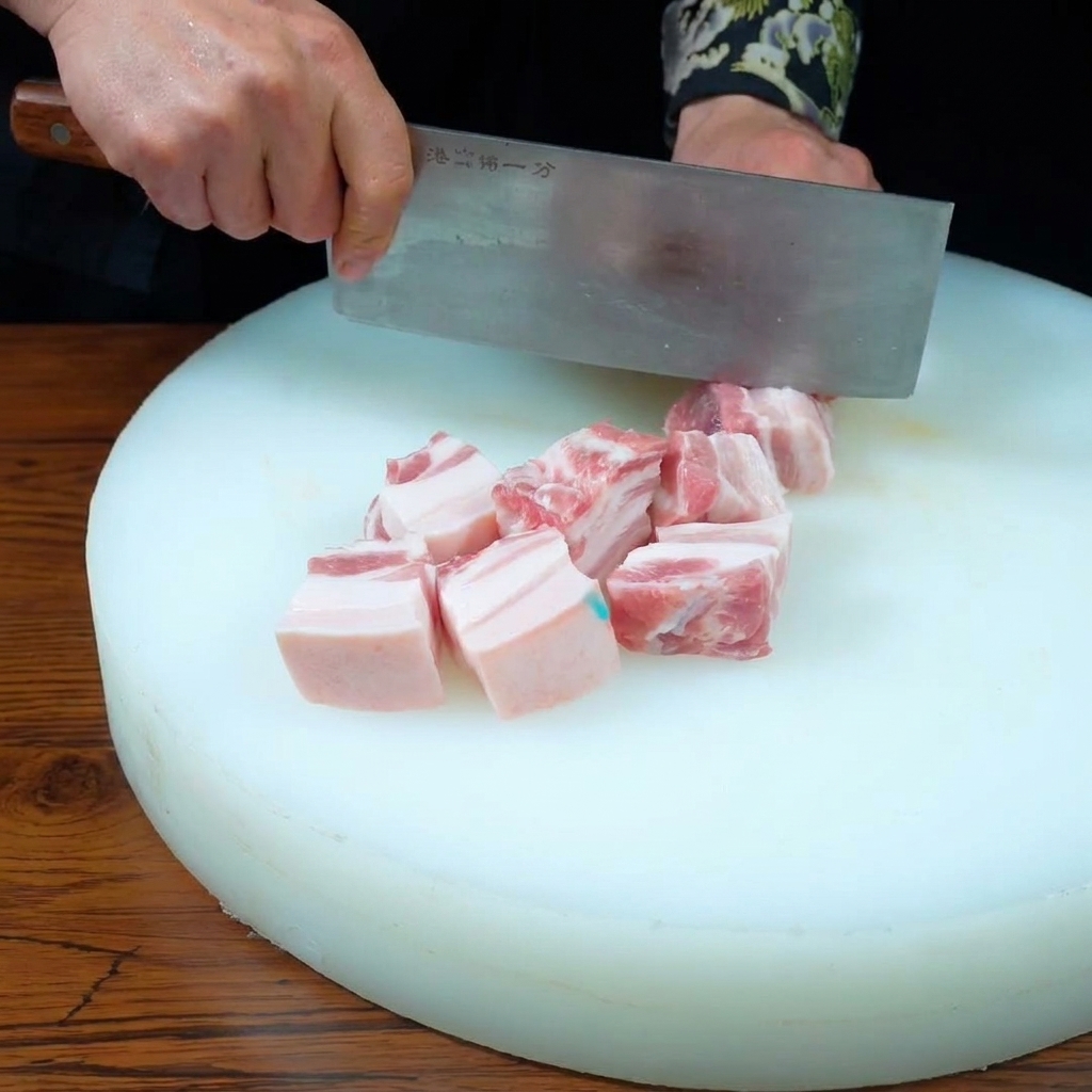 Mani che usano una mannaia per tagliare un trancio di pancia di maiale a tre strati in cubetti uniformi su un tagliere bianco.