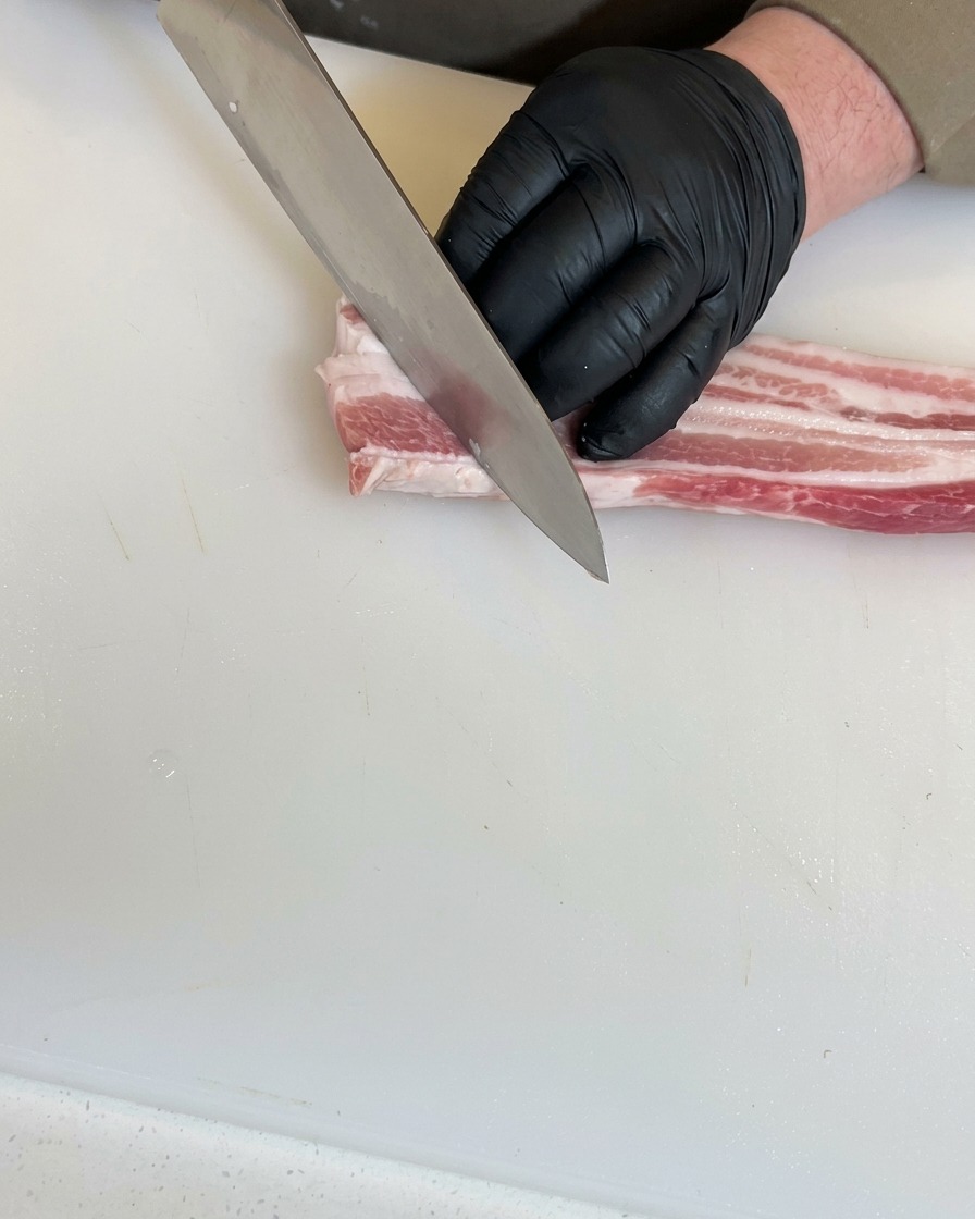 Mani con guanti neri usano un coltello da chef per praticare tagli diagonali profondi in un pezzo di pancetta cruda.