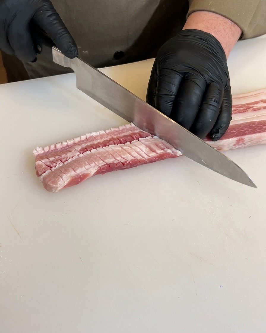 Mani con guanti neri incidono il lato opposto della pancetta creando una trama a griglia.