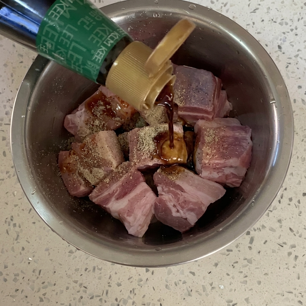 Salsa di soia versata da una bottiglia sui cubetti di pancetta conditi in una ciotola di metallo.