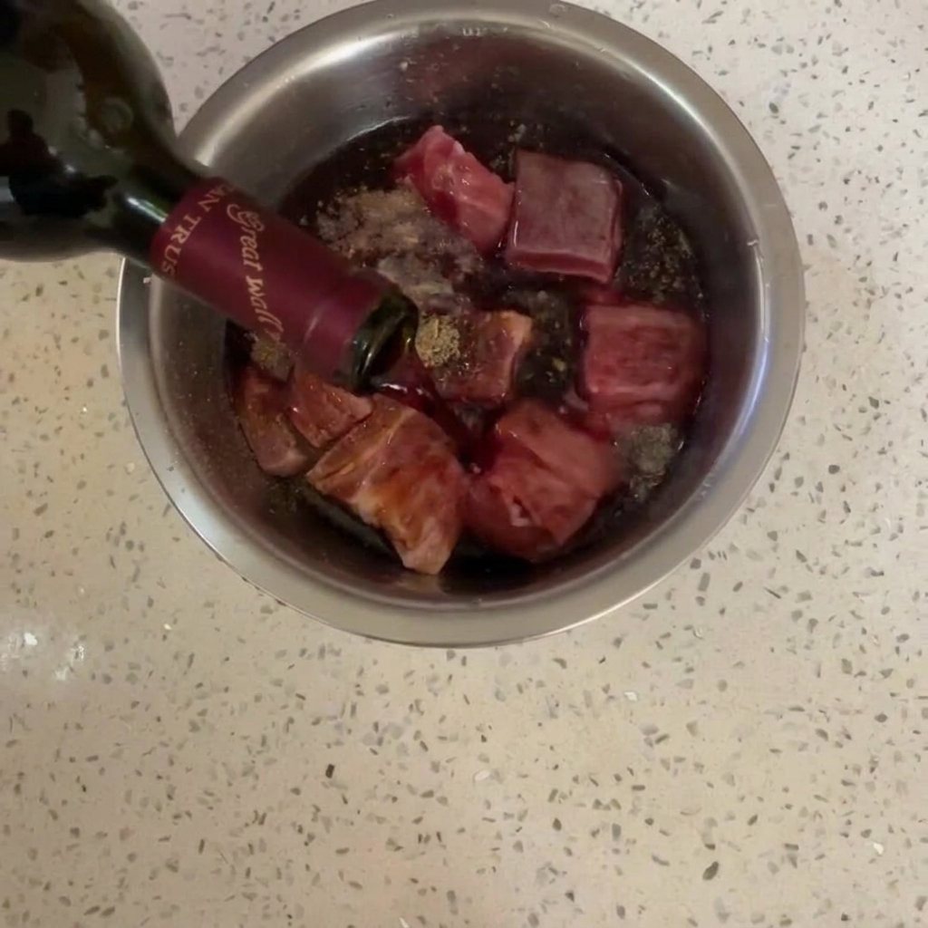 Vino rosso versato da una bottiglia di vetro scuro sulla pancetta condita in una ciotola di acciaio inox.