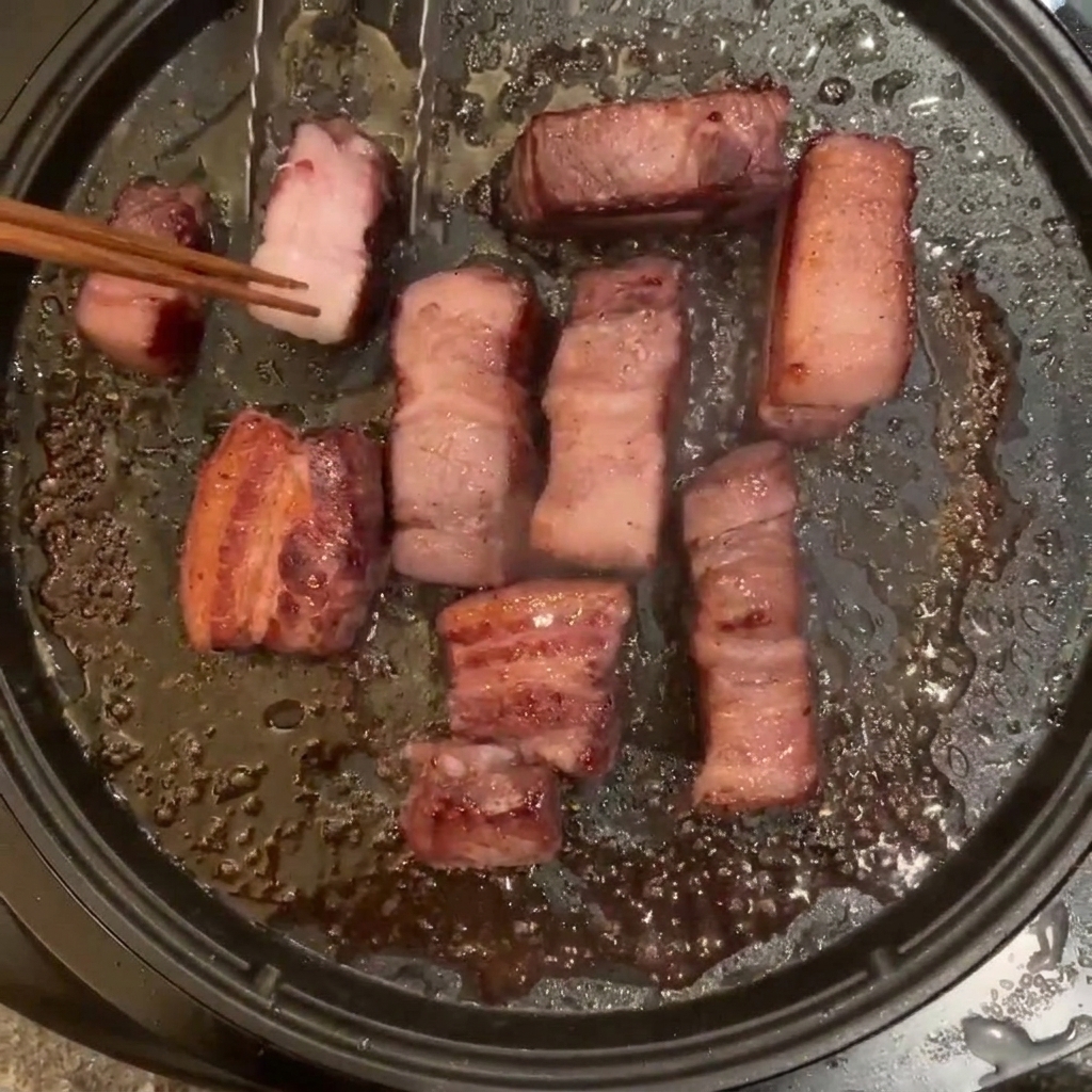 Diversi pezzi di pancetta di maiale che friggono in una padella elettrica, girati con bacchette di legno.