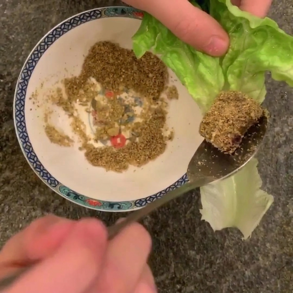 Primo piano di un cucchiaio con un pezzo di pancetta cotta ricoperta di arachidi tritate, sopra una foglia di lattuga romana.