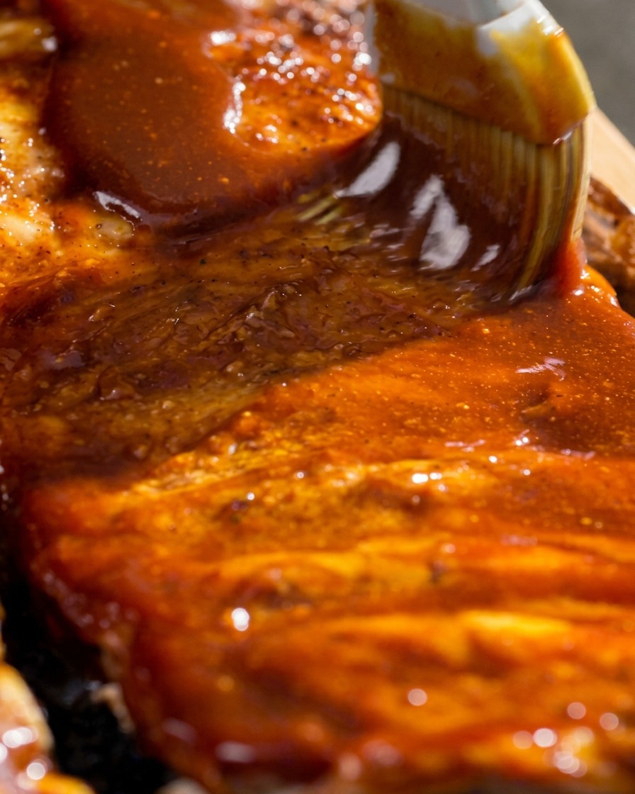 Primo piano di un pennello da cucina che stende una glassa barbecue rossa e lucida sulle costine cotte.