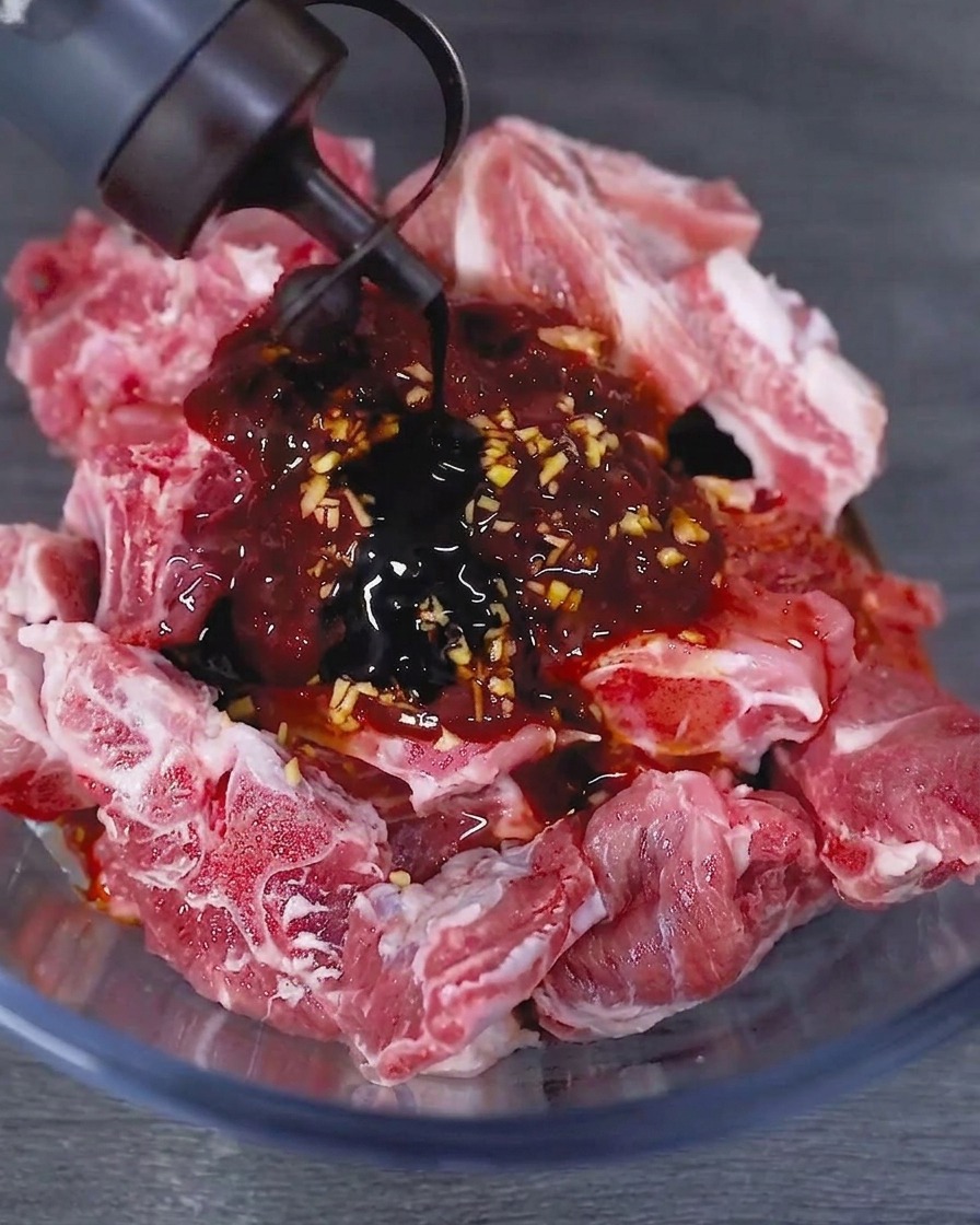 Salsa di soia scura versata da una bottiglia in una ciotola di vetro contenente le costine di maiale crude marinate.