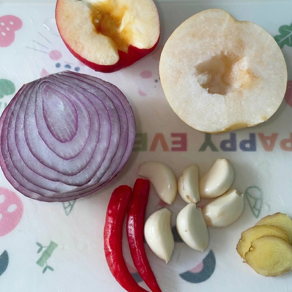 Mela, mezza cipolla rossa, mezza pera, spicchi daglio, peperoncini rossi e zenzero su un tagliere.