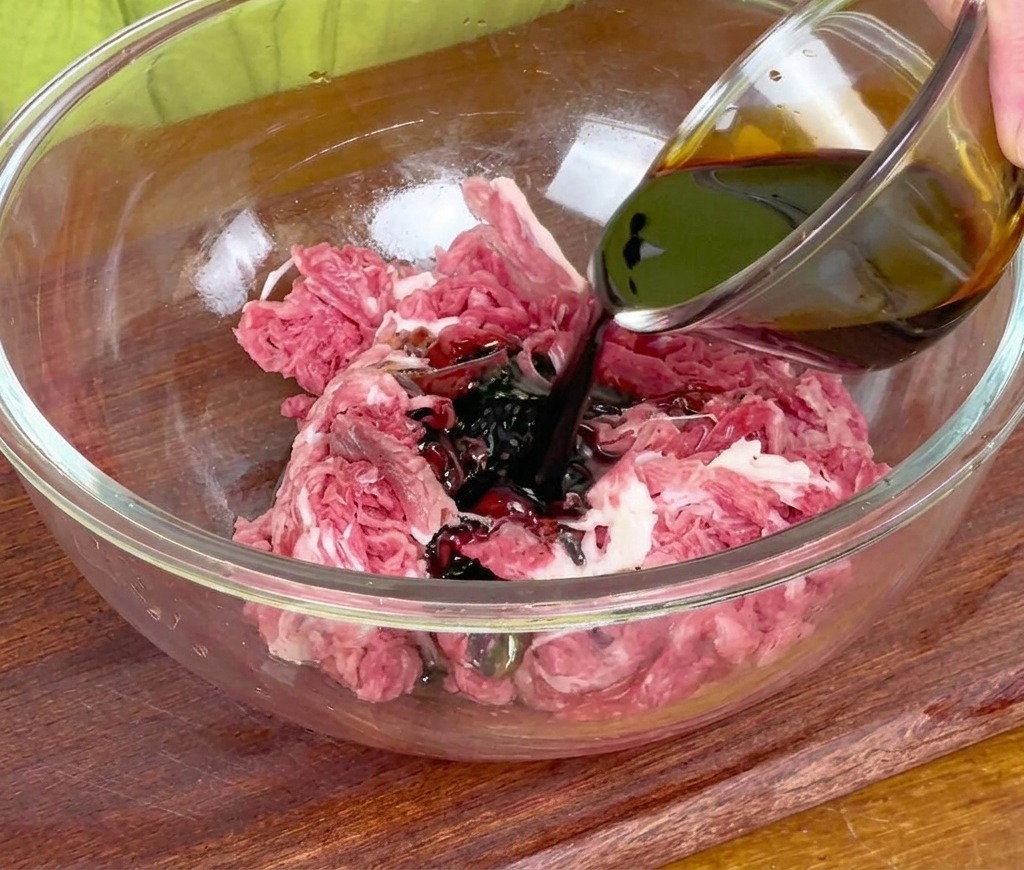 Versare la salsa di soia scura da una piccola ciotola di vetro sul manzo crudo affettato in una grande ciotola di miscelazione.