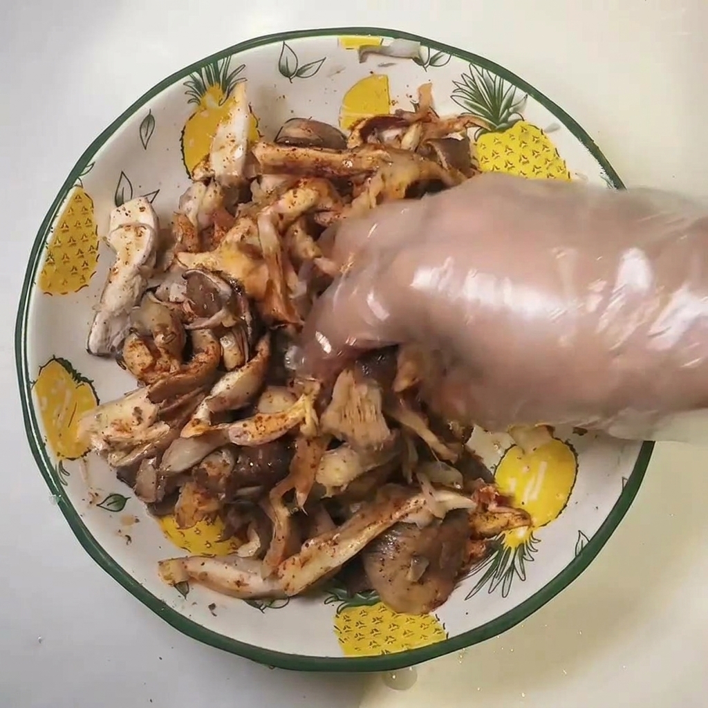 Una mano con guanto di plastica che mescola i funghi conditi in una ciotola.