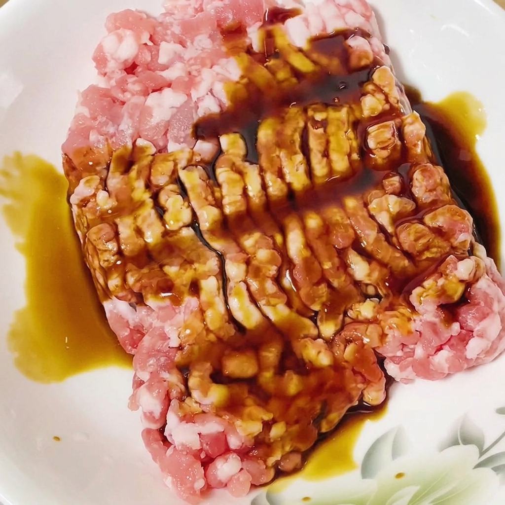 Salsa di soia versata sul maiale macinato crudo in una ciotola bianca.
