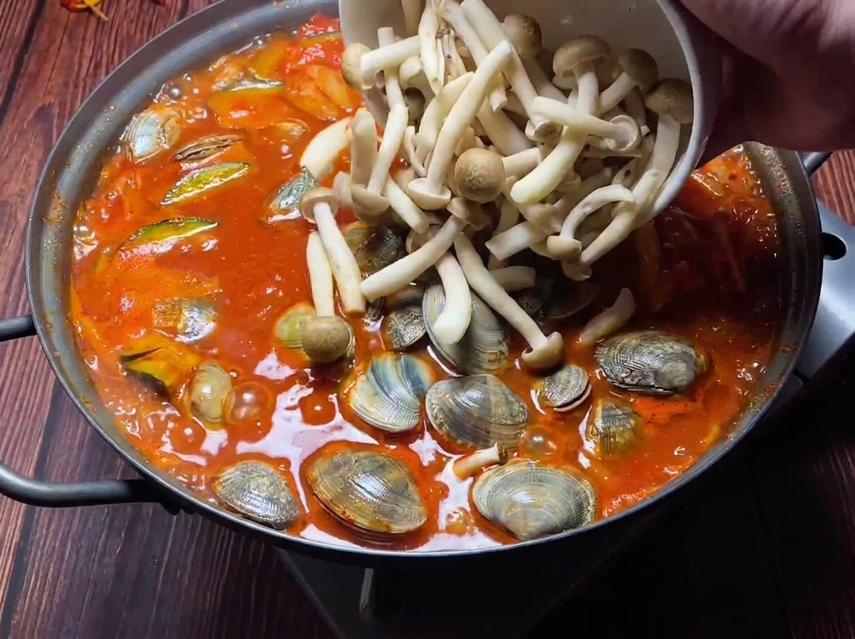 Vongole fresche e funghi misti aggiunti a uno stufato di kimchi rosso bollente.