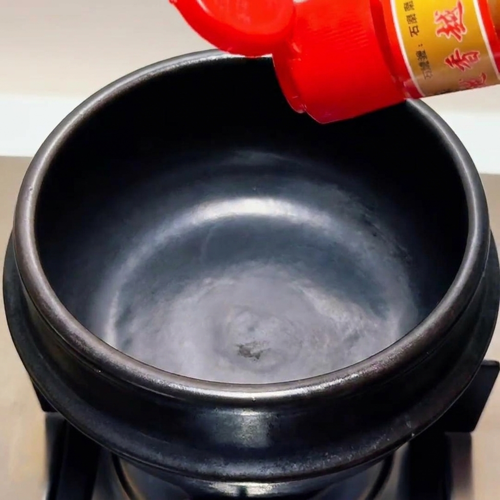 Olio di sesamo versato da una bottiglia con tappo rosso in un pentolino di pietra nero vuoto.