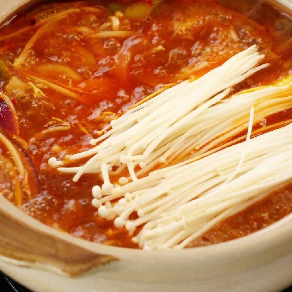 Funghi enoki freschi posizionati in un brodo rosso piccante che sobbolle nella pentola.