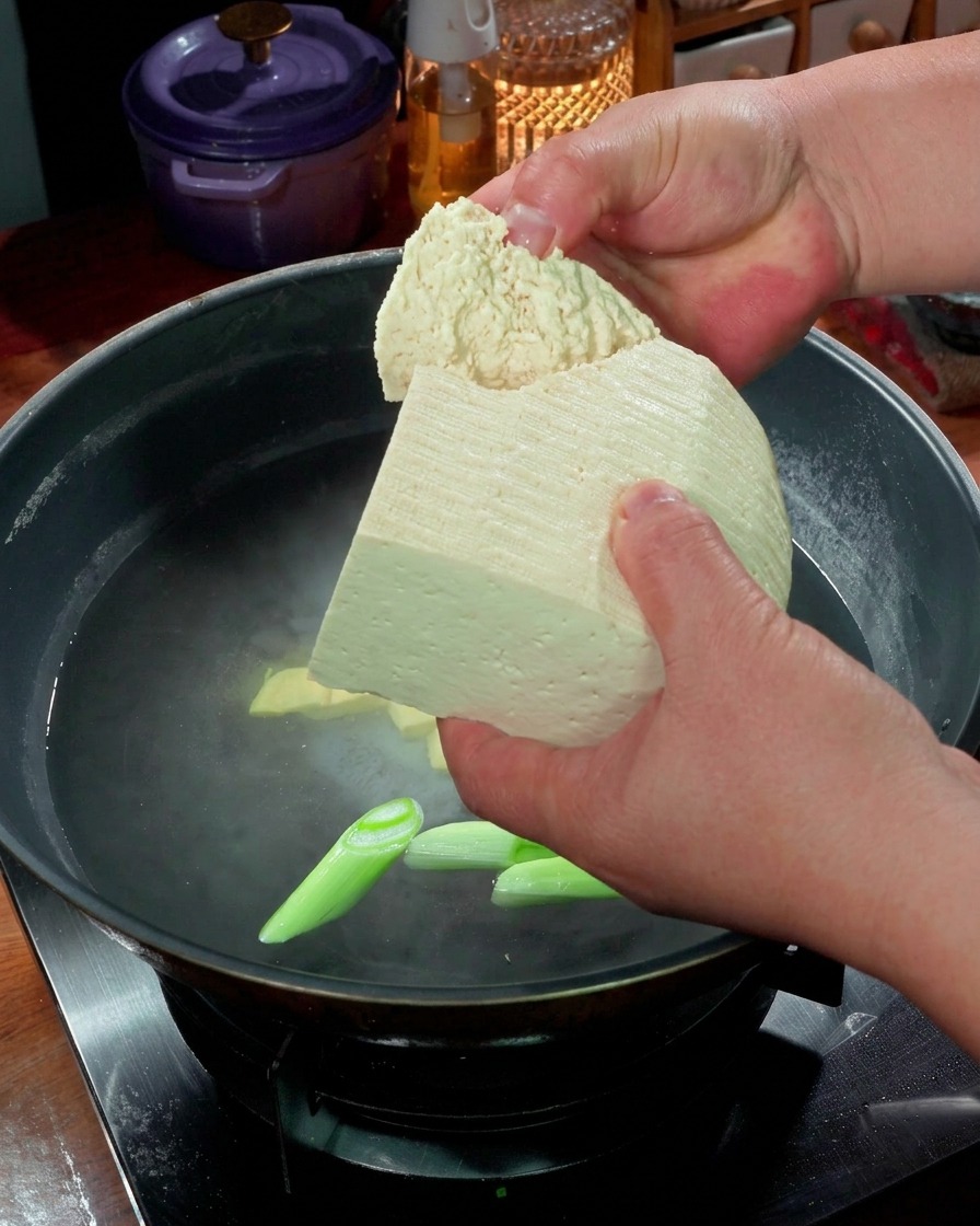 Mani che rompono pezzi di tofu compatto in una pentola di acqua bollente con cipollotti.