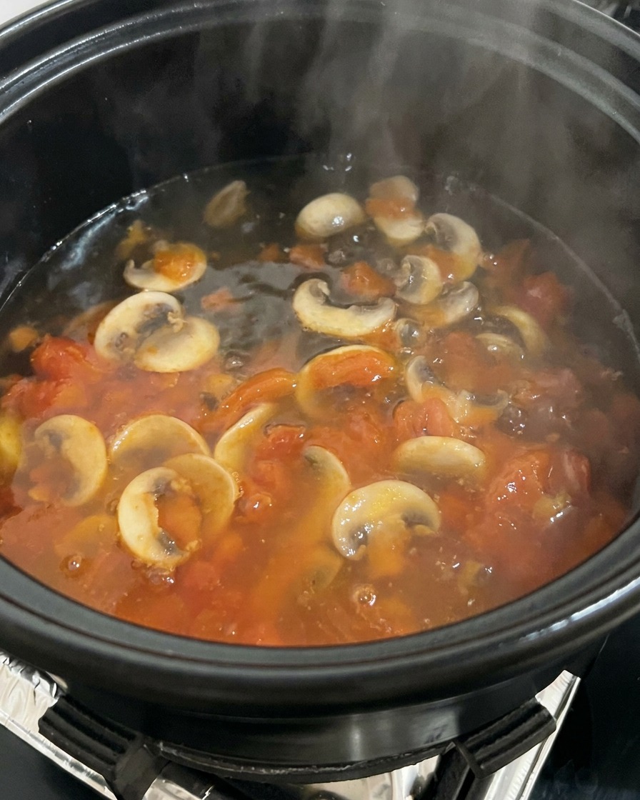Acqua bollente versata in una pentola piena di pomodori e funghi saltati, creando vapore.