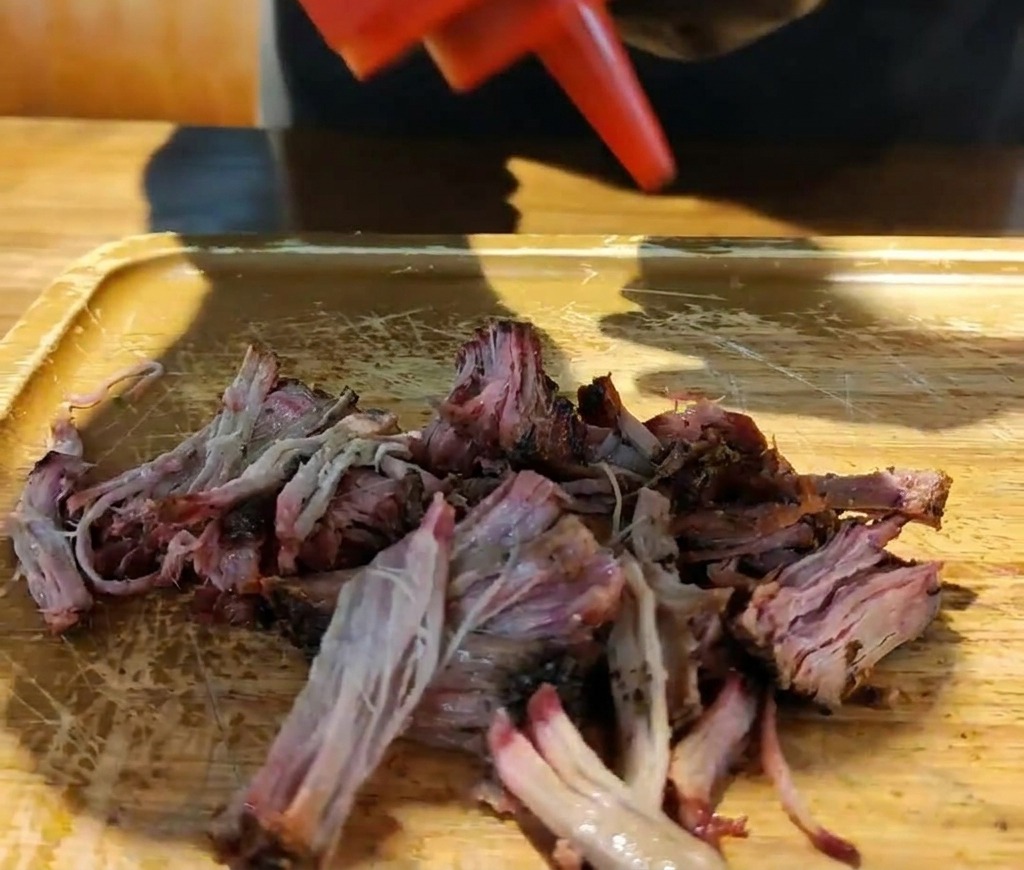 Salsa barbecue densa versata da una bottiglia sopra il maiale nero sfilacciato sul tagliere.
