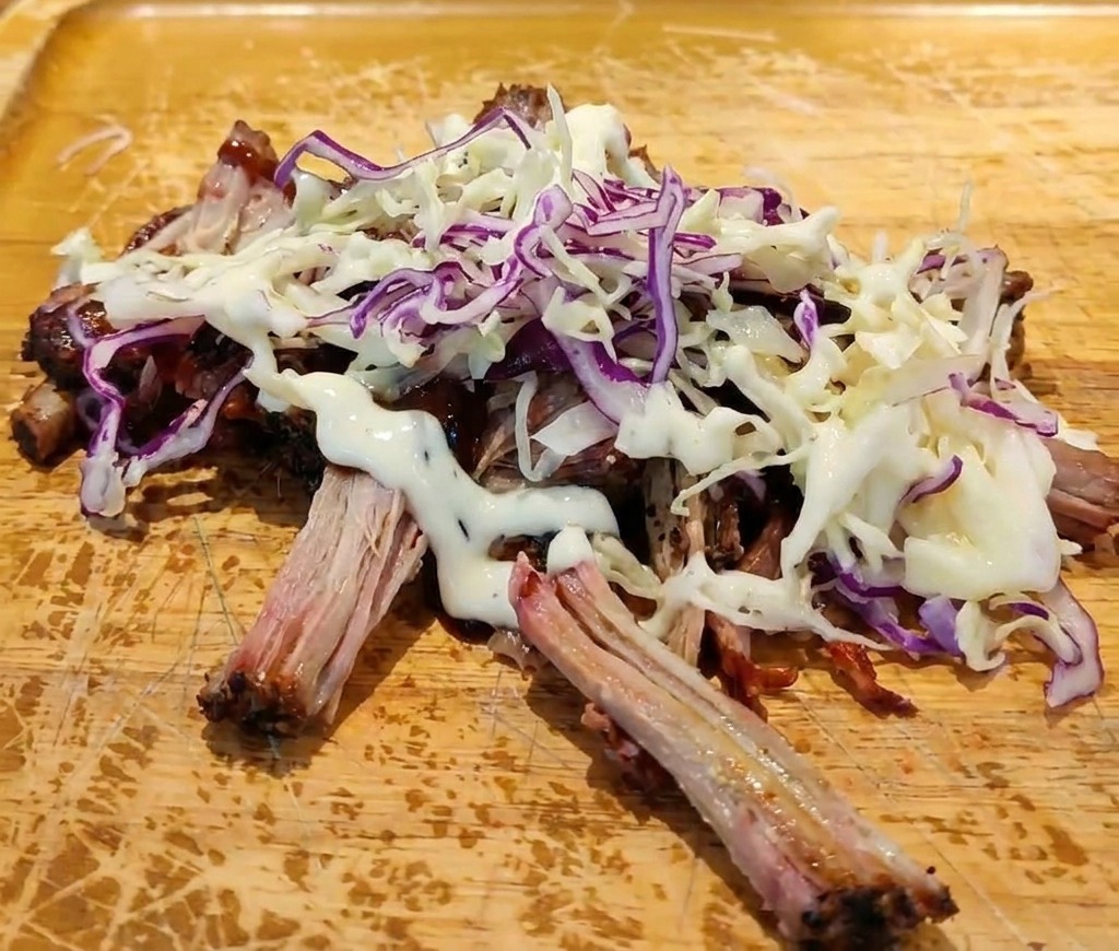 Maiale sfilacciato con salsa BBQ sormontato da coleslaw e un filo di salsa bianca cremosa su un tagliere.
