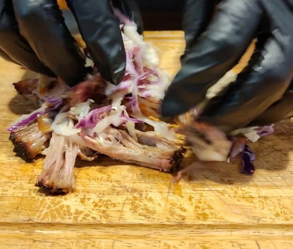 Mani esperte che mescolano maiale, salsa scura e coleslaw colorato su una superficie di legno.