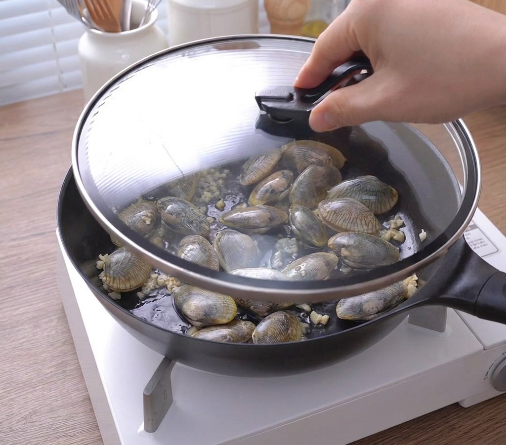 Una mano mette un coperchio di vetro sopra una padella piena di vongole e aglio.