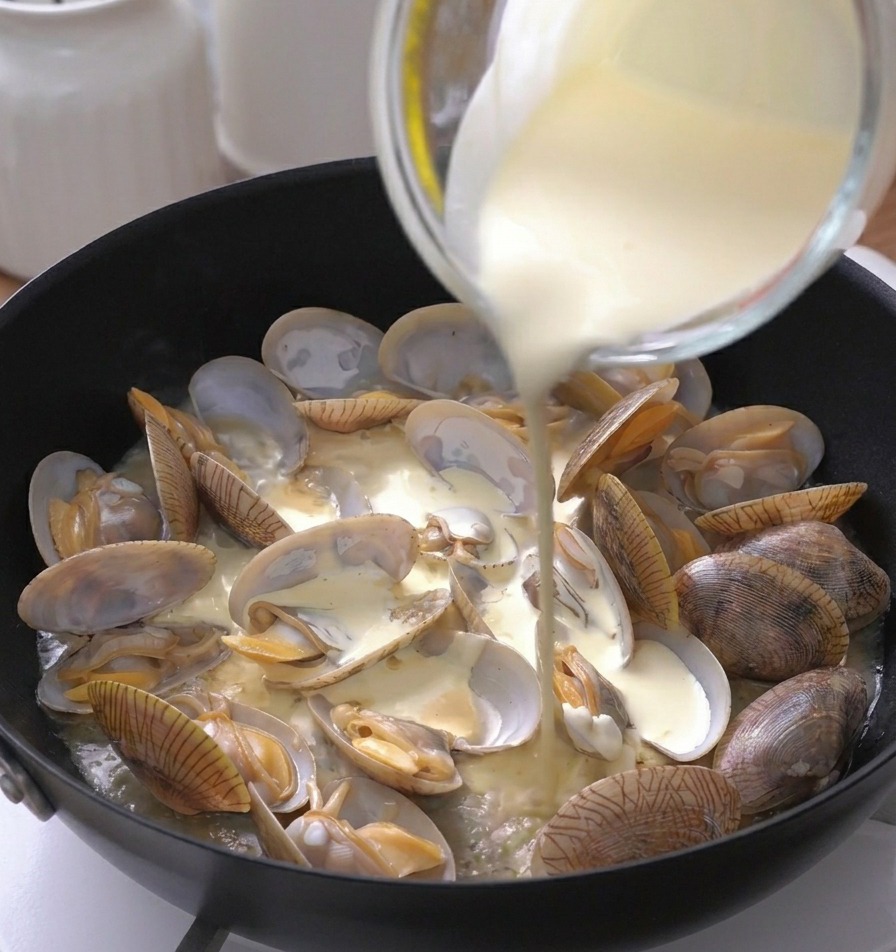 Panna liquida bianca versata da un misurino in una padella con vongole aperte.