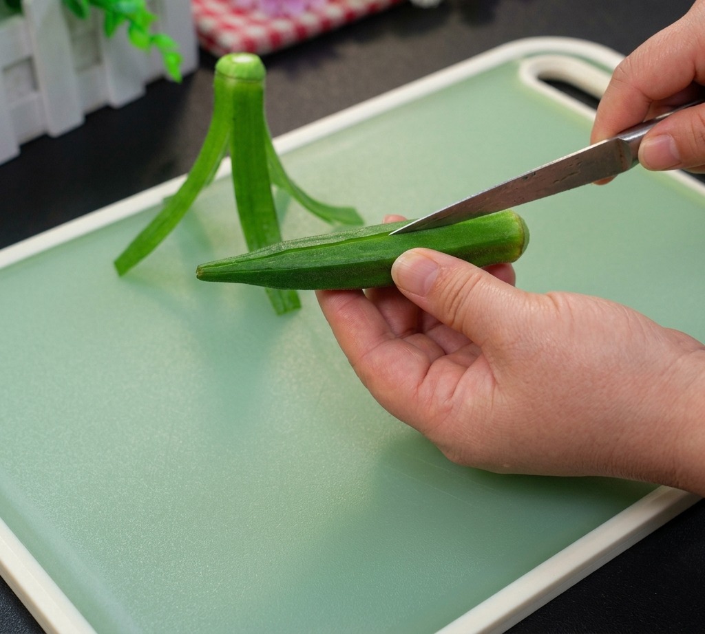 Una mano che tiene un baccello di okra verde intero su un tagliere mentre un coltello inizia a tagliare il bordo superiore.