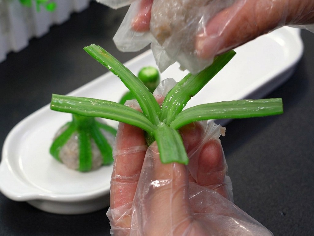 Una persona con guanti in plastica che posiziona una pallina di macinato di gamberi rosato al centro delle strisce di okra sbollentate.
