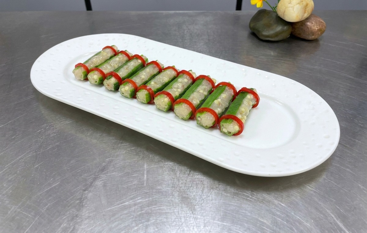 Due file ordinate di okra farcita con anelli di peperoncino su un piatto ovale bianco, pronte per la cottura.