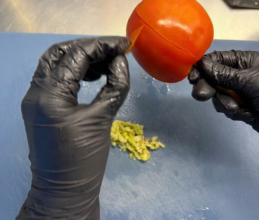 Mani con guanti neri rimuovono la sottile buccia esterna da un pomodoro rosso maturo su un tagliere.