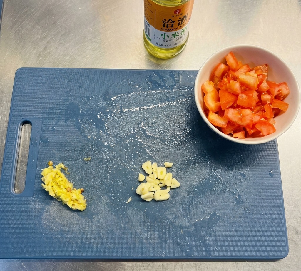 Un tagliere blu mostra aglio affettato, peperoncini tritati e pomodori a cubetti accanto a una bottiglia dolio.