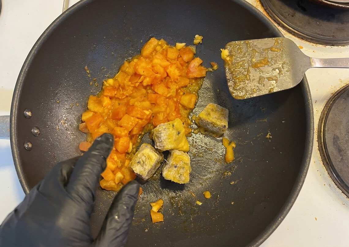 Cubetti di polpa di frutto della passione e peperoncini tritati aggiunti ai pomodori saltati da una mano con guanto nero.