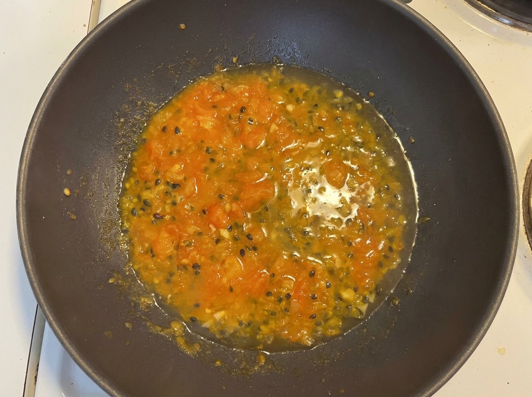 Acqua aggiunta al mix di pomodoro e frutto della passione in un wok, creando una base arancione con semi di frutto della passione visibili.