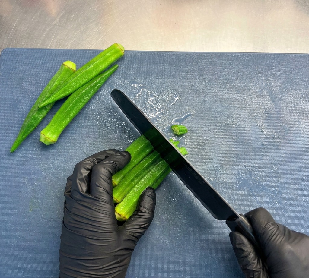 Una persona affetta i gambi di okra verde brillante in piccole rondelle circolari su un tagliere blu.