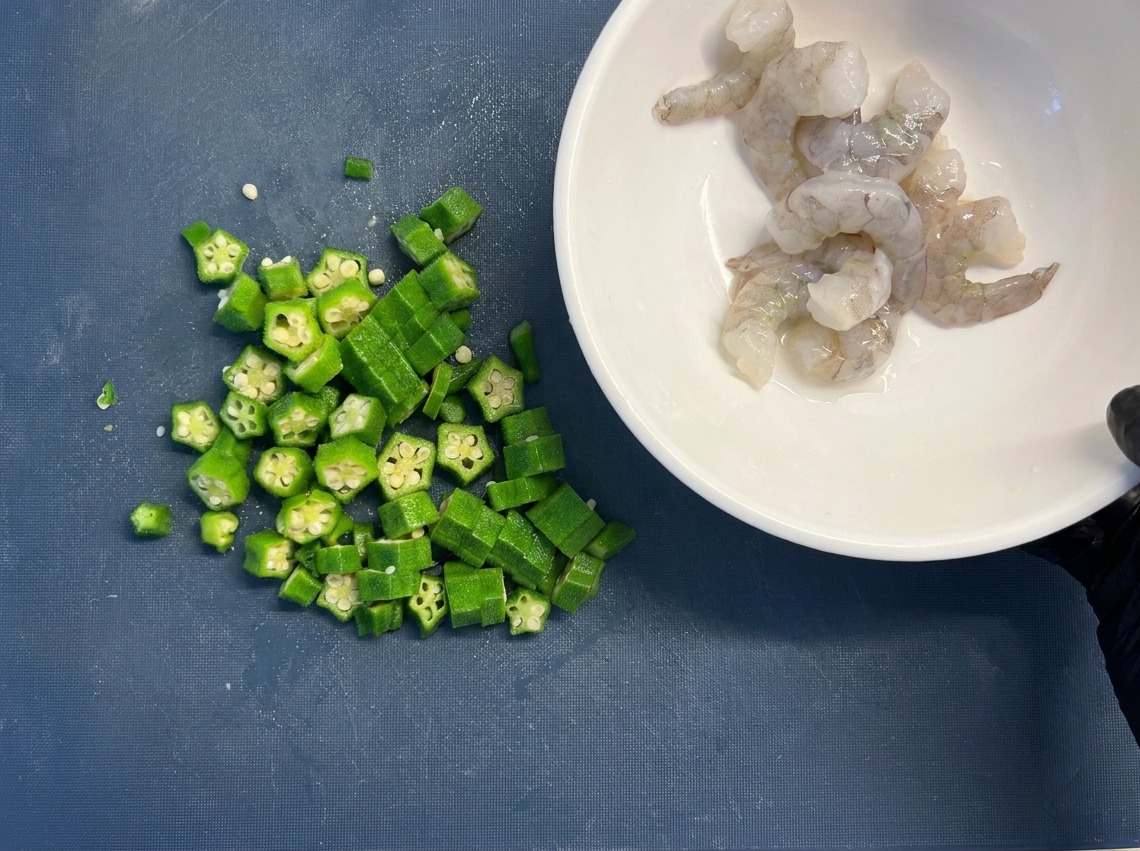 Una ciotola bianca piena di gamberi crudi puliti tenuta accanto a una pila di rondelle di okra su un tagliere blu.