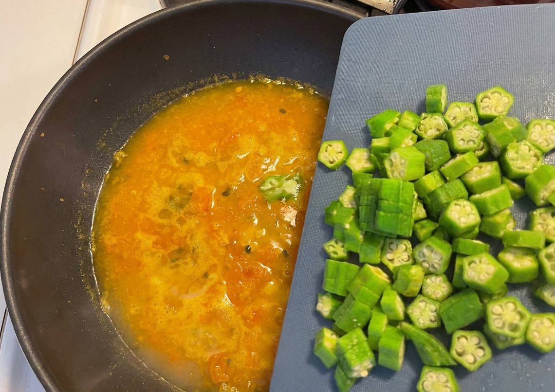 Rondelle di okra fresca versate da un tagliere blu in una zuppa arancione che bolle in un wok scuro.