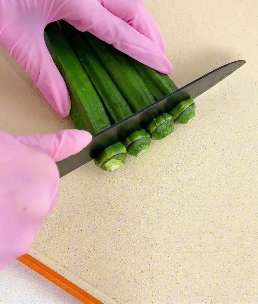 Mani con guanti rosa che tagliano le estremità di un mazzetto di okra verde con un coltello nero.