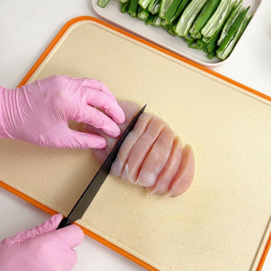 Petto di pollo crudo affettato con un coltello nero su un tagliere chiaro da mani con guanti rosa.