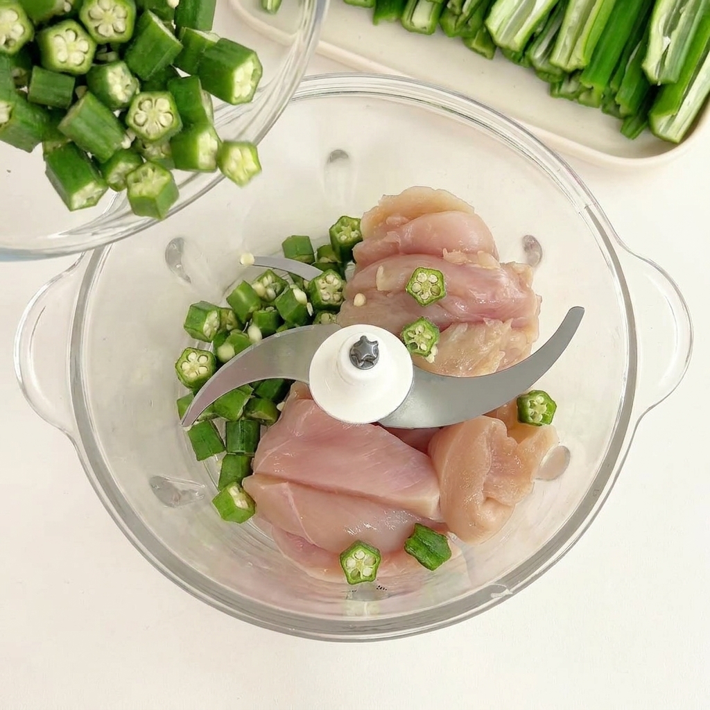 Semi di okra verde versati da una ciotolina su pezzi di pollo crudo in un robot da cucina.