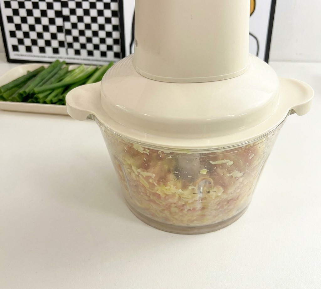 Un robot da cucina color crema su un piano bianco, riempito con un trito fine di carne di pollo e semi di okra.