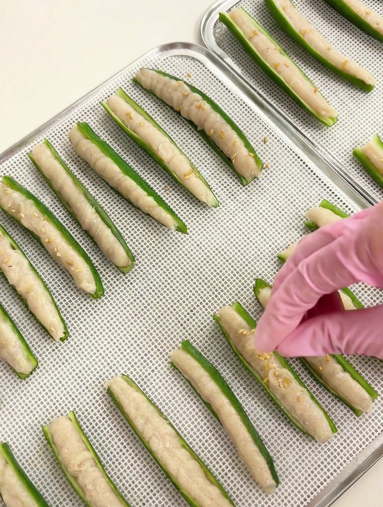 Dettaglio di una mano che sparge semi di sesamo bianco su file di okra farcite su un vassoio argentato.