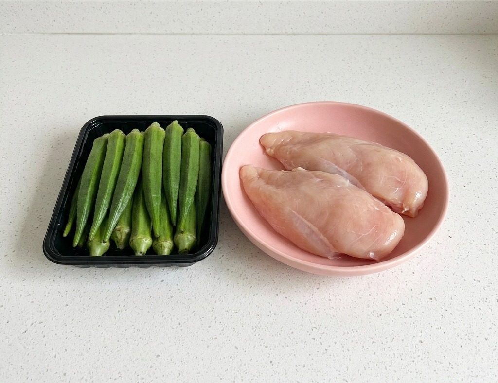Un vassoio nero con baccelli di okra verdi freschi accanto a un piatto con due petti di pollo crudi su un piano di lavoro bianco.