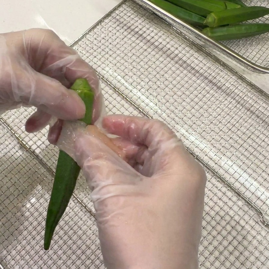 Mani che avvolgono una sottile fetta di pollo a spirale intorno a un intero baccello di okra verde.
