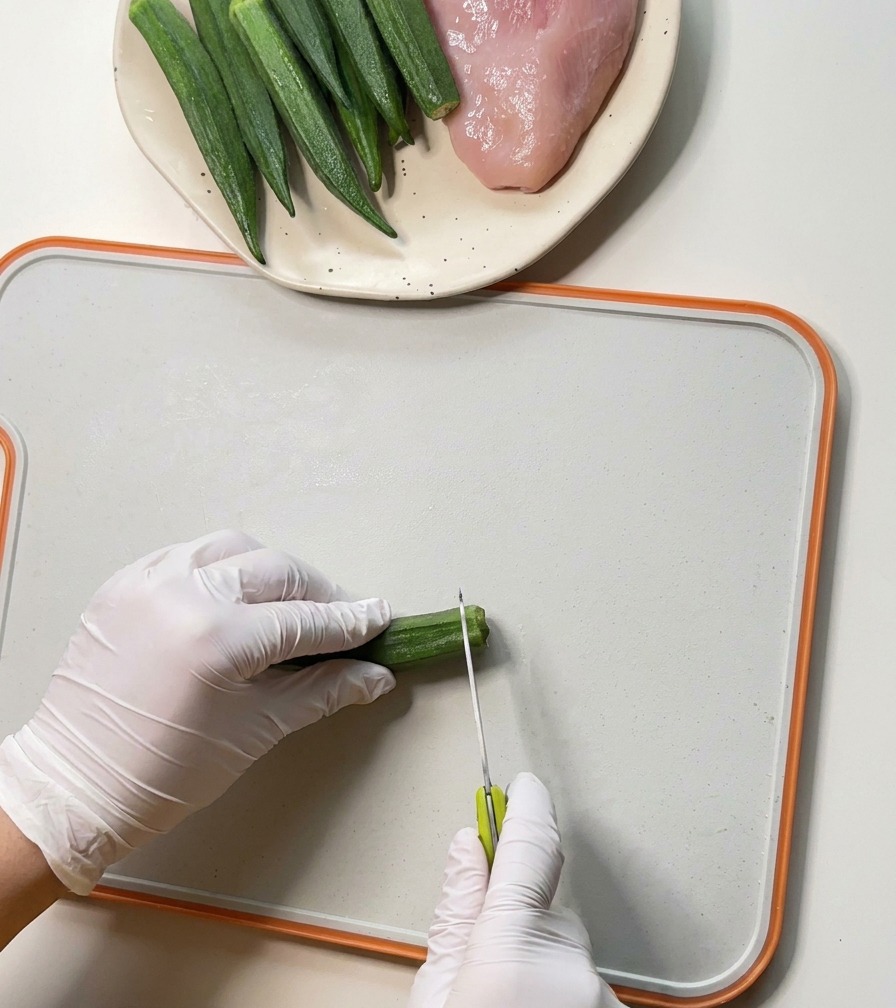 Mani con guanti che usano uno spelucchino per rimuovere le estremità di baccelli di okra freschi su un tagliere bianco.