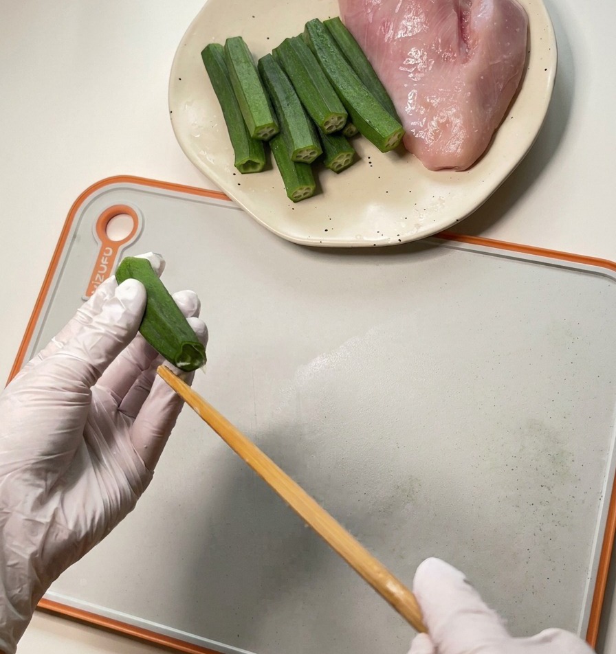 Una persona che indossa guanti bianchi usa una bacchetta di legno per estrarre semi e polpa da un baccello di okra verde.