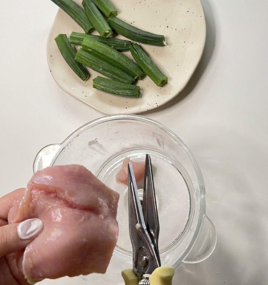 Mani che usano forbici da cucina in metallo per tagliare il petto di pollo crudo direttamente in un boccale del frullatore.