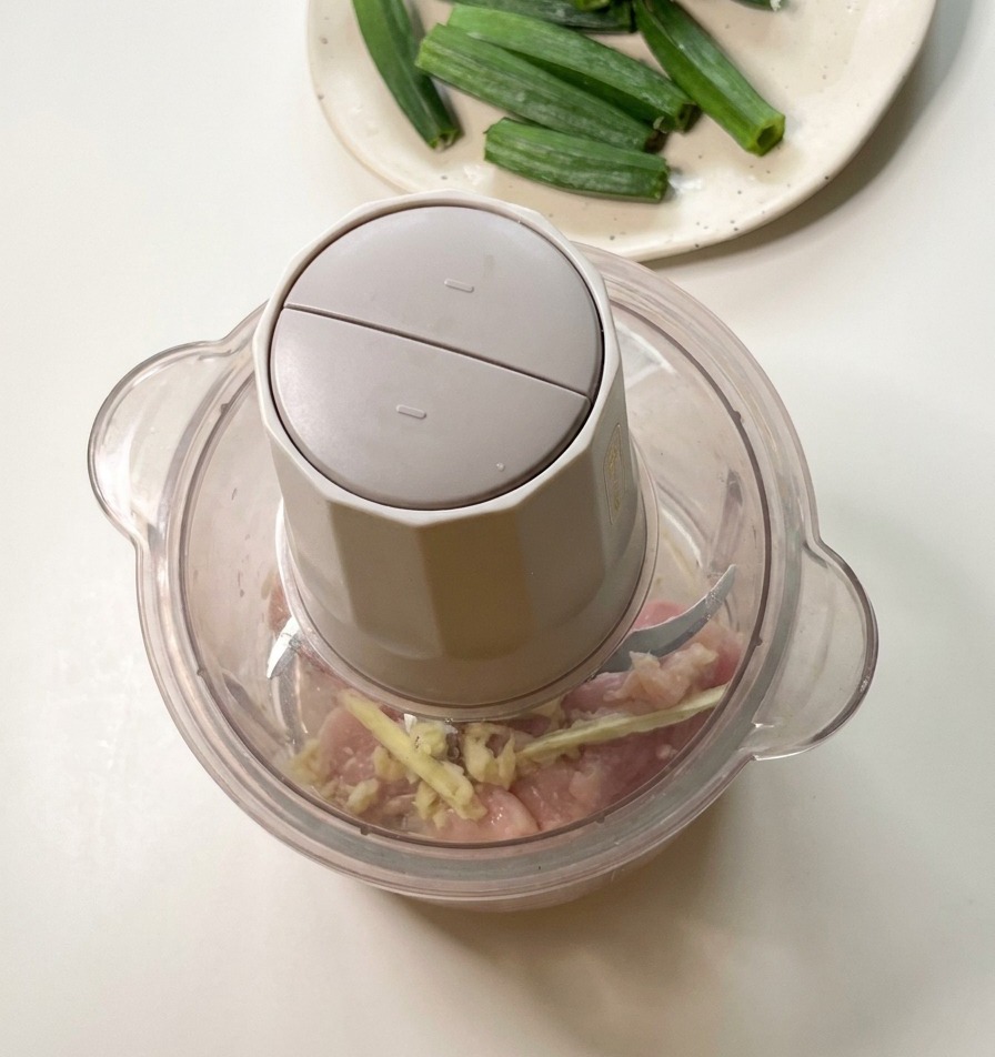Un robot da cucina su un bancone contenente pezzi di pollo crudo e semi di okra, pronto per la lavorazione.