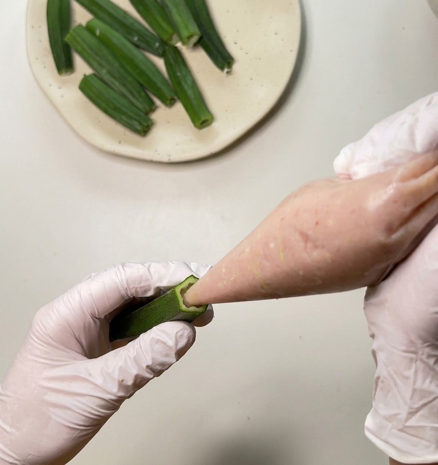 Primo piano di una persona che versa la pasta di pollo rosa in un baccello di okra verde cavo usando una sac à poche.