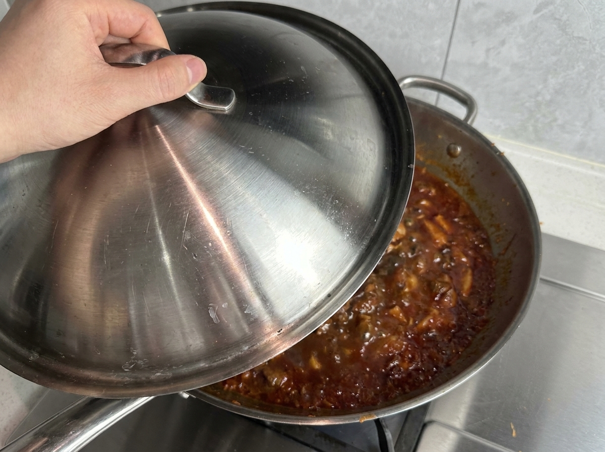 Una mano mette un coperchio dacciaio su una grande padella tipo wok piena di sugo di carne rosso che gorgoglia.