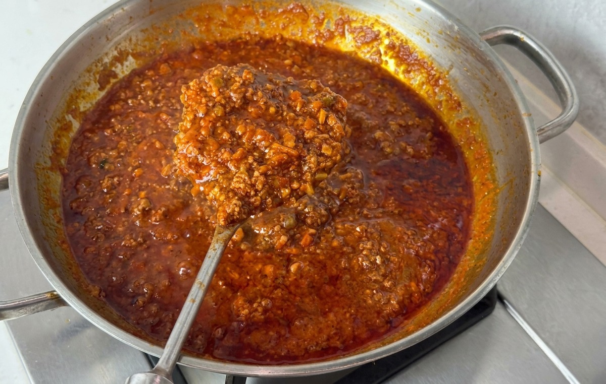 Una padella dacciaio con un ragù denso e scuro sollevato da un grande cucchiaio di metallo per mostrare la sua consistenza viscosa.