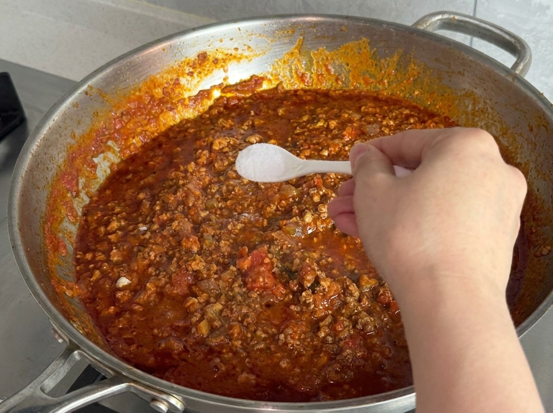 Una mano tiene un cucchiaino di plastica bianco pieno di sale sopra una padella con sugo di carne rosso che bolle.